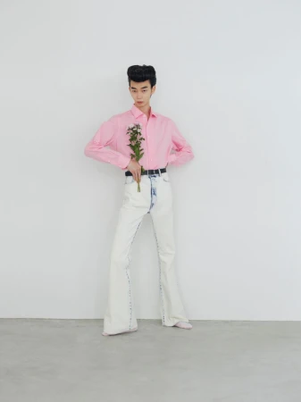 Model wearing a pink long-sleeve shirt and white pants holding a small bouquet of flowers against a seamless backdrop