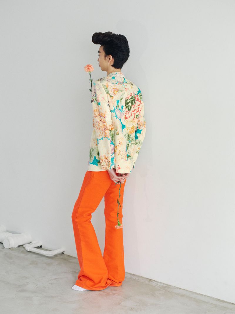 Model with styled dark hair holding a flower wearing a floral print jacket and bright orange trousers
