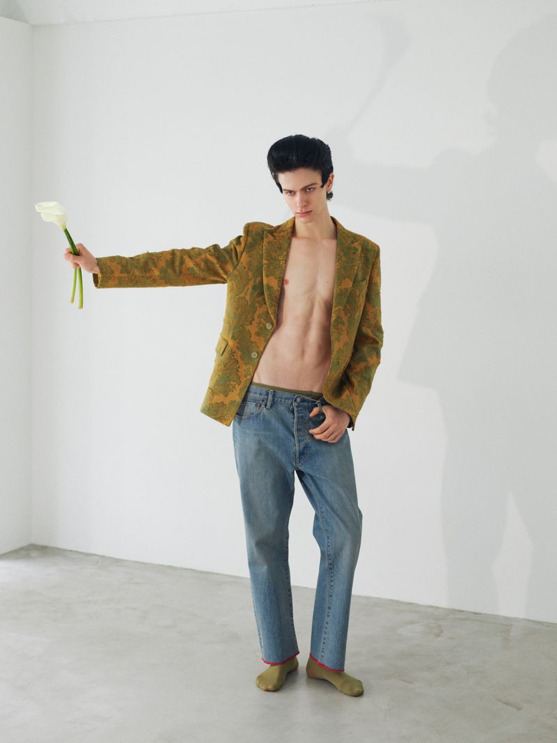 A shirtless male model wearing an olive patterned blazer and blue jeans holds a calla lily against a white studio wall.