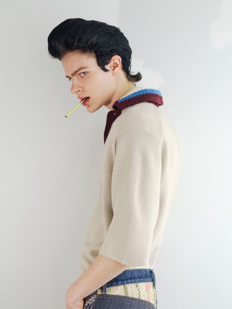 Profile portrait of a young male model with styled dark hair holding a cigarette in his mouth, wearing a collared knit top.