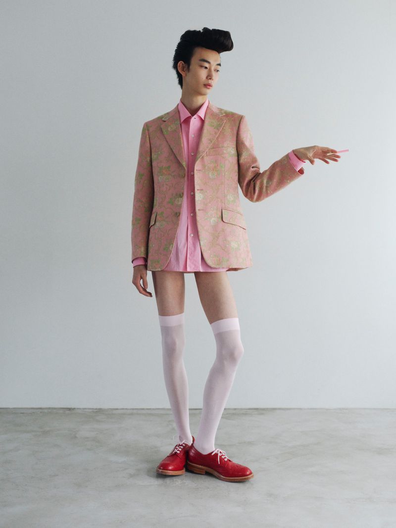 Model with styled dark hair wearing pink patterned blazer, pink shirt, knee-high socks, and red shoes standing against a grey wall