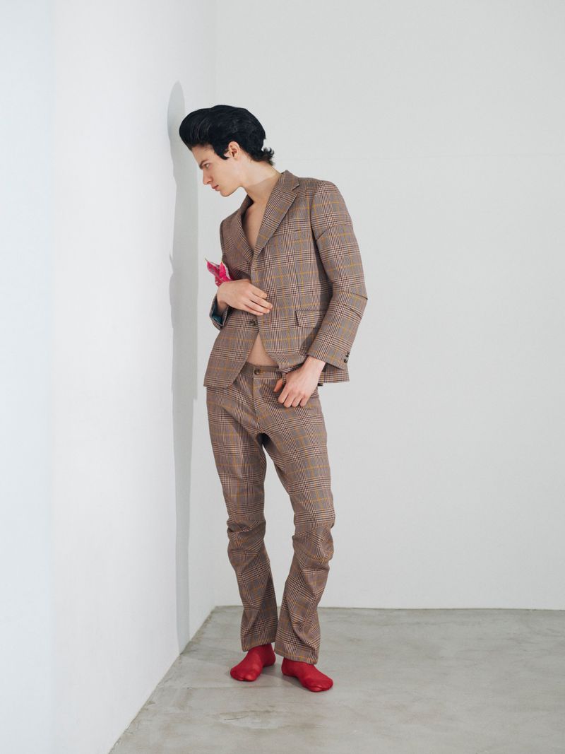 Man wearing a brown plaid suit and red shoes leaning against a plain white wall