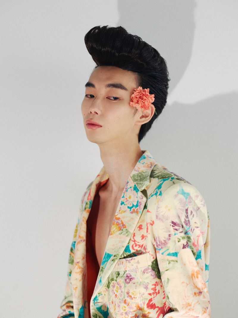 East Asian man with voluminous pompadour hair and floral jacket featuring a pink flower tucked behind his ear against a plain white wall