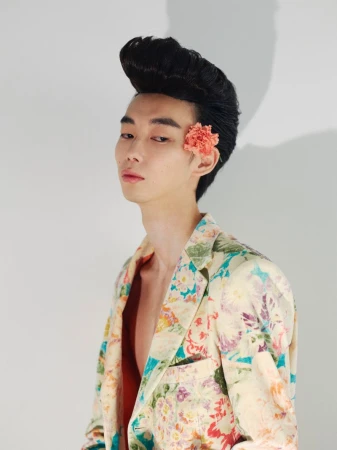 East Asian man with voluminous pompadour hair and floral jacket featuring a pink flower tucked behind his ear against a plain white wall