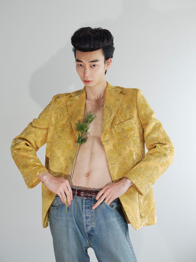 Model in a patterned yellow jacket and blue jeans holding a green stem against a plain white background.