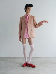 Model with styled dark hair wearing pink patterned blazer, pink shirt, knee-high socks, and red shoes standing against a grey wall