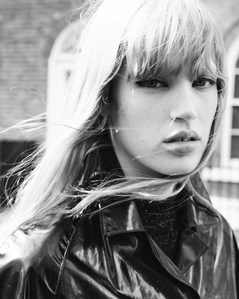 Close-up black and white portrait of a young adult woman with blonde hair and a black vinyl coat, shot outdoors against a brick wall.