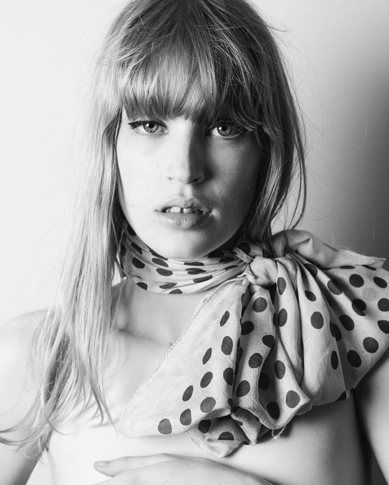 Black and white portrait of a young woman with bangs and a polka dot scarf, posing with hand near chest, close-up view, indoor setting.