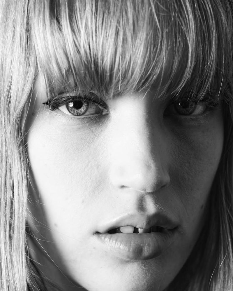 Close-up black and white portrait of a blonde woman with straight hair, visible eyeliner, fair skin, and slight underbite. Studio shot.