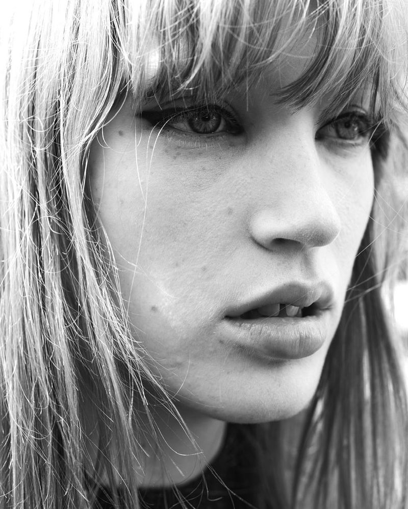 Striking monochrome portrait of a young adult female with bangs and winged eyeliner, gazing slightly off-camera, against a soft background.