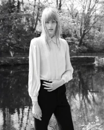 Model Jacqui Hooper in a white long-sleeve blouse and black trousers outdoors by a river in a black and white portrait.