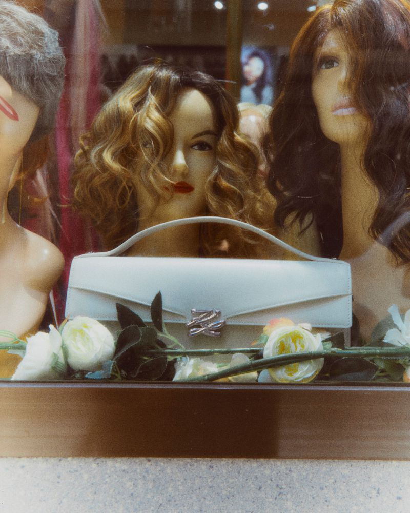 Mannequin heads displaying wigs behind a glass display case with a white handbag and floral decorations for ODDA Magazine Karl Lagerfeld project.