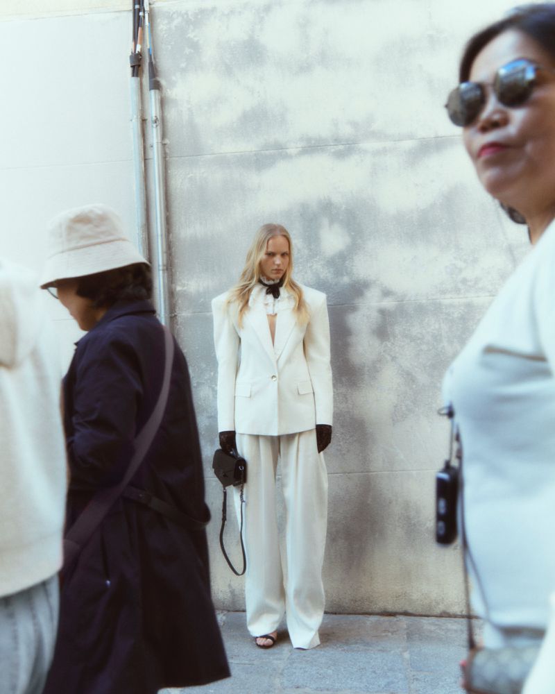 Model in white suit standing against an urban wall with figures in the foreground