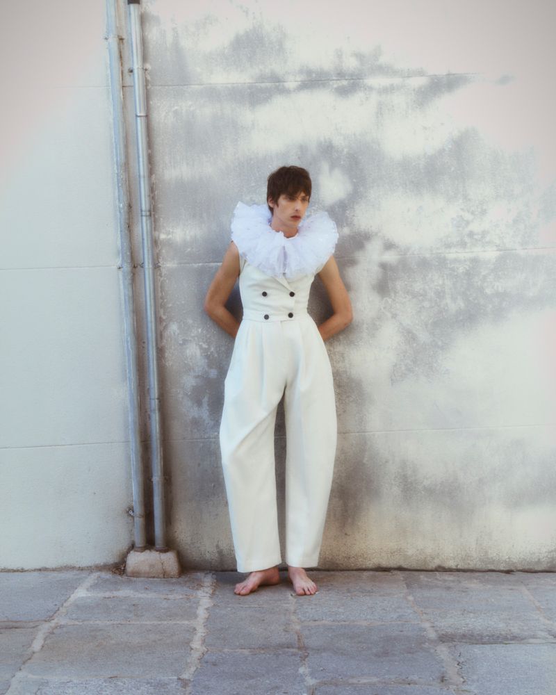 Model wearing a white jumpsuit with a ruffled collar standing barefoot against a weathered concrete wall for ODDA Magazine.