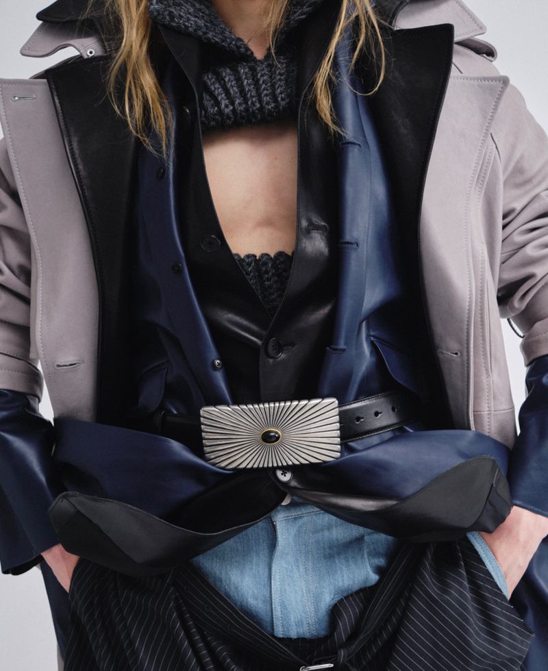 Close-up portrait of a model wearing layered dark clothing with a prominent silver buckle belt and light trench coat against a plain backdrop.
