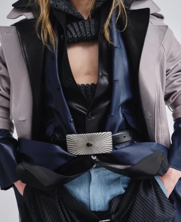 Close-up portrait of a model wearing layered dark clothing with a prominent silver buckle belt and light trench coat against a plain backdrop.