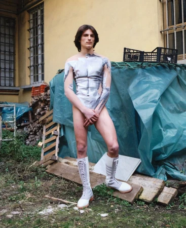A slim adult male poses in body paint and tall boots in an outdoor setting with a building and tarpaulin in the background.