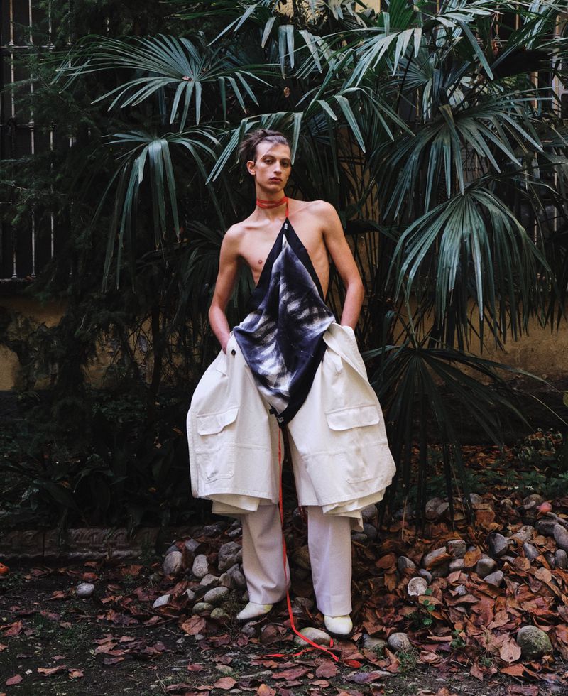 Model standing outdoors wearing a draped patterned top and white cargo trousers against lush tropical foliage.