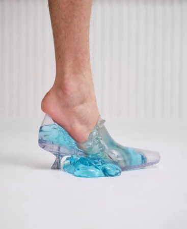 Bare lower leg wearing a transparent blue liquid-filled high heel shoe against a white studio seamless background.