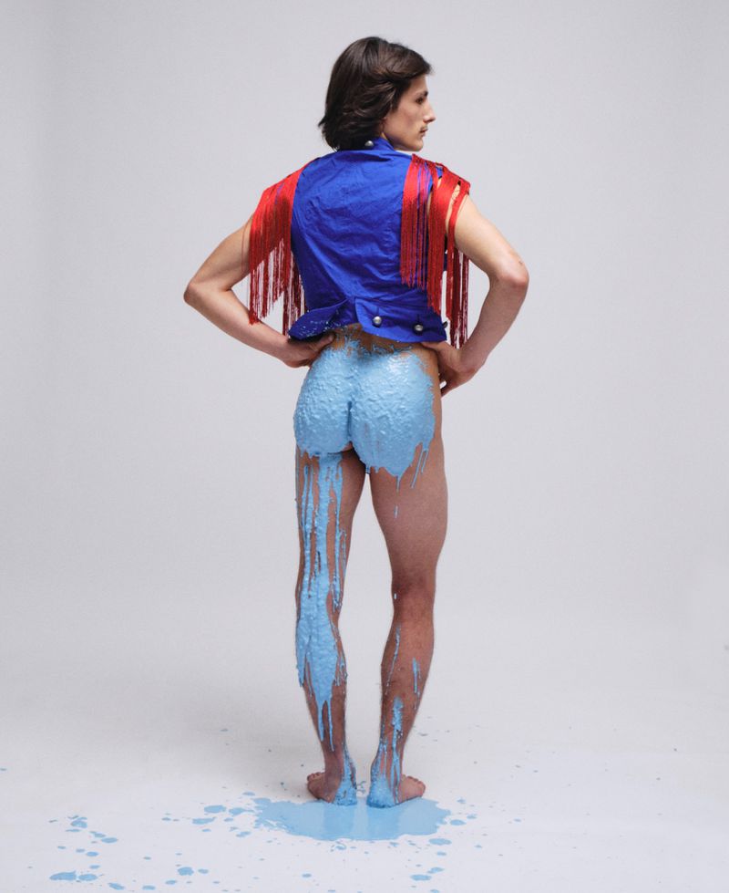 Model from behind wearing a blue vest with red fringe, with blue paint dripping down his legs against a white background