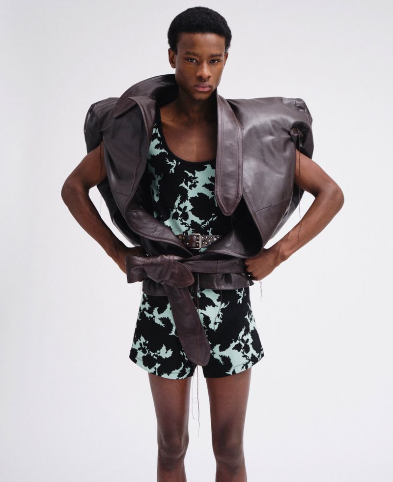 Model wearing an oversized brown leather jacket and patterned outfit against a plain white studio background.