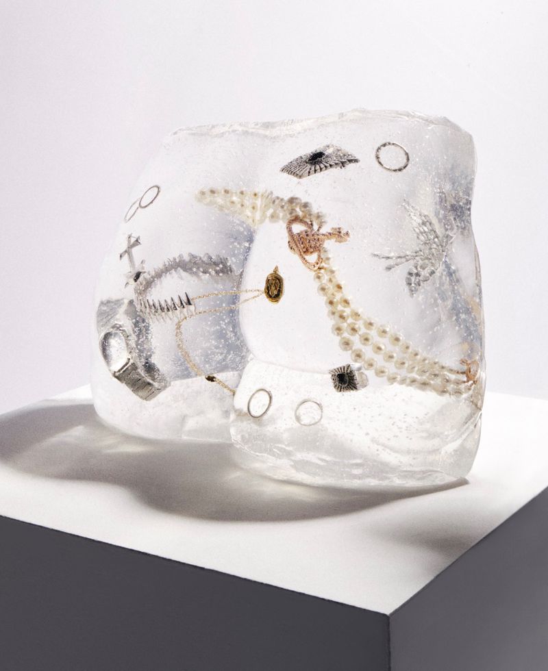 A block of ice containing various pieces of jewellery displayed on a white pedestal in a studio setting.