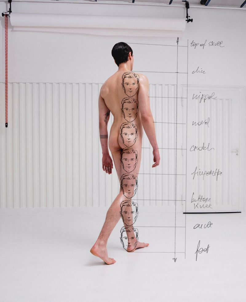 Back view of a bare-chested man walking, with male faces drawn on his back and leg, posing in a white photo studio.