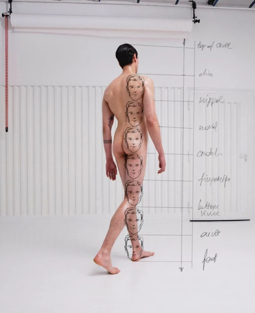 Back view of a bare-chested man walking, with male faces drawn on his back and leg, posing in a white photo studio.