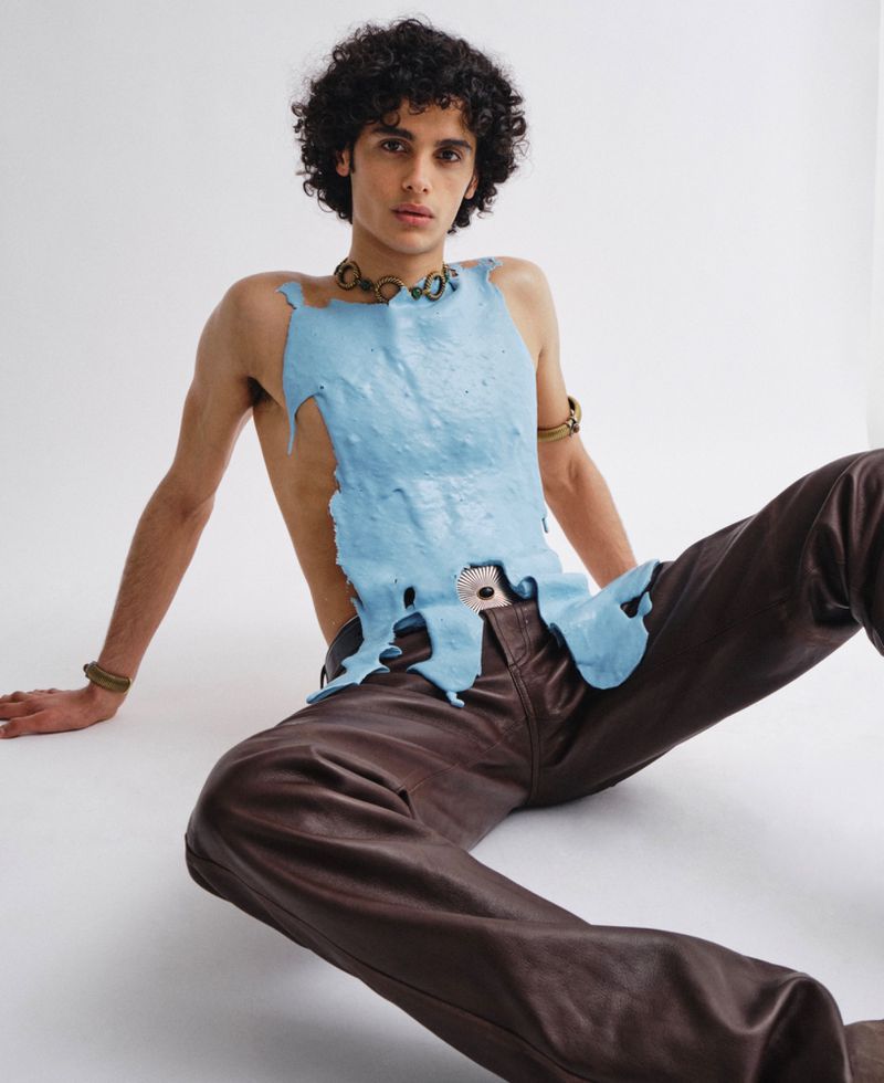 Model with dark curly hair wearing a blue textured top and brown leather trousers seated against a white background.