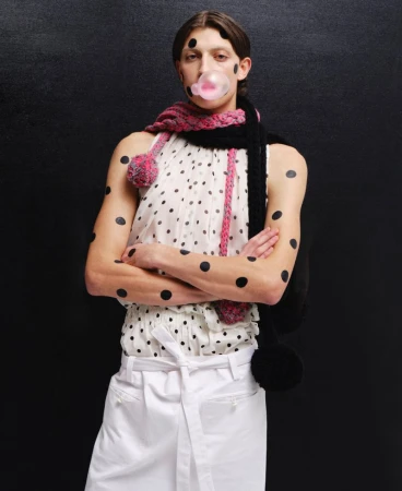 Model blowing a pink bubblegum bubble wearing a polka dot top and knitted scarf against a dark background