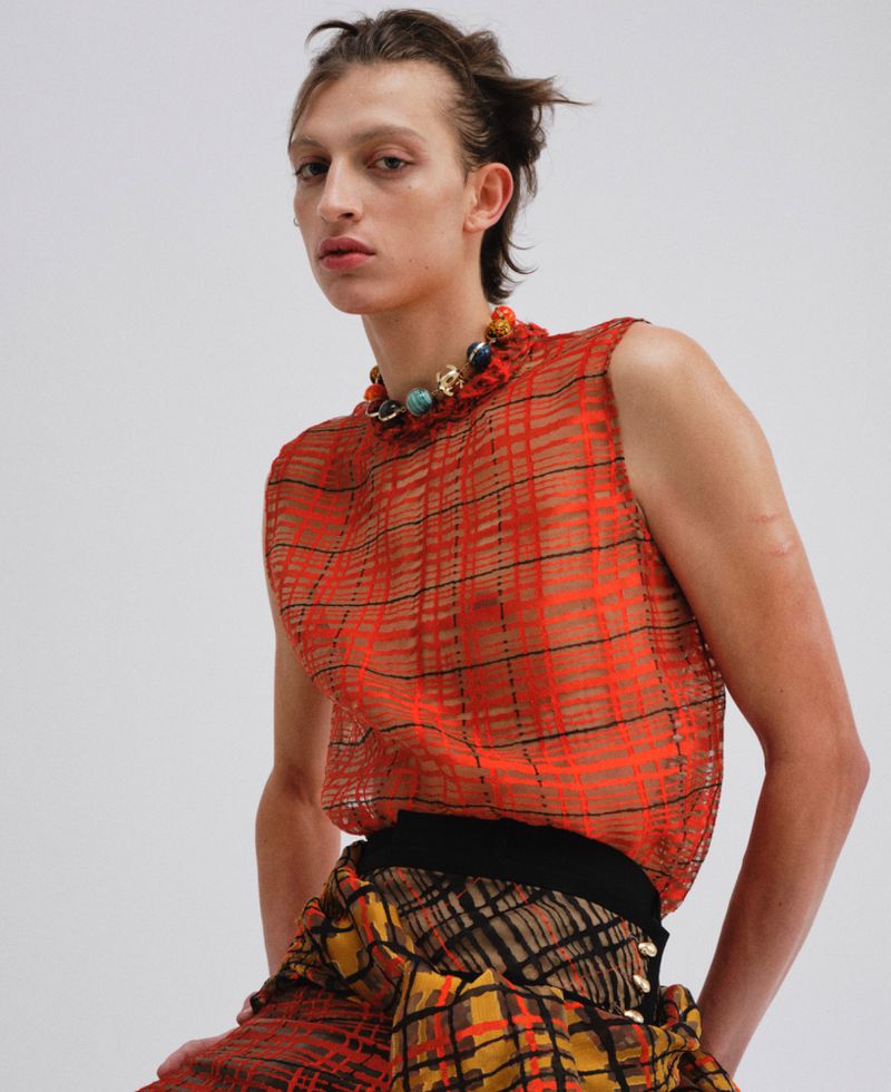 Model wearing a sheer red plaid sleeveless top and beaded necklace against a plain white studio background.
