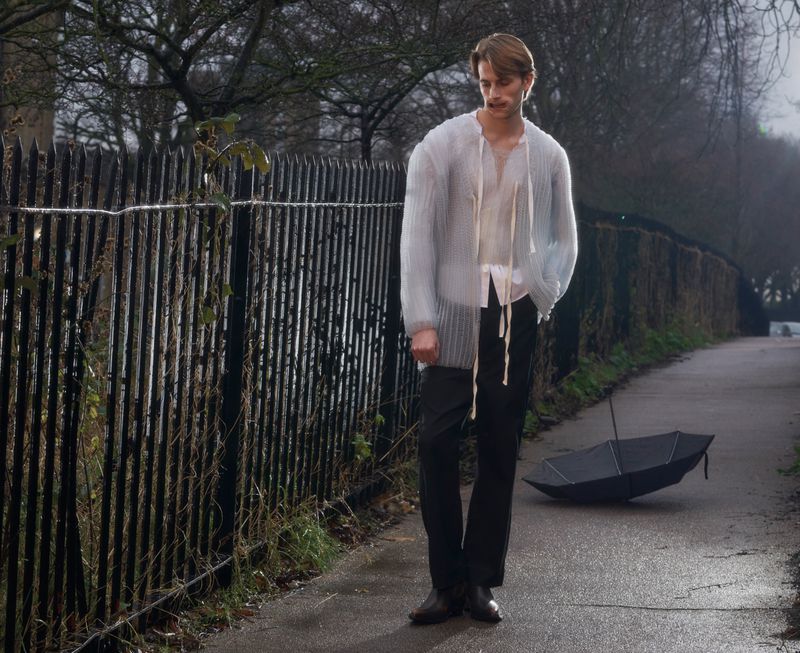 A young male model wearing an open sheer knit cardigan and dark trousers stands near a metal fence with an umbrella on the path.
