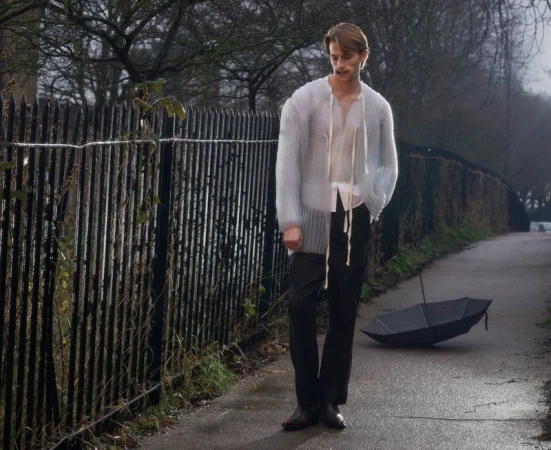 A young male model wearing an open sheer knit cardigan and dark trousers stands near a metal fence with an umbrella on the path.