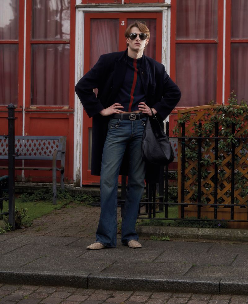 Model wearing dark coat, blue jeans, and sunglasses standing outside a red building with a bench and fence.
