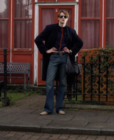 Model wearing dark coat, blue jeans, and sunglasses standing outside a red building with a bench and fence.