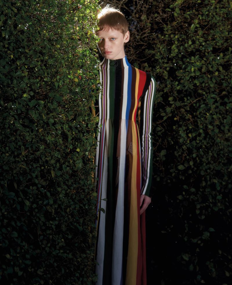 Model wearing a vertical striped multi-color dress standing among dark green foliage under dappled sunlight.