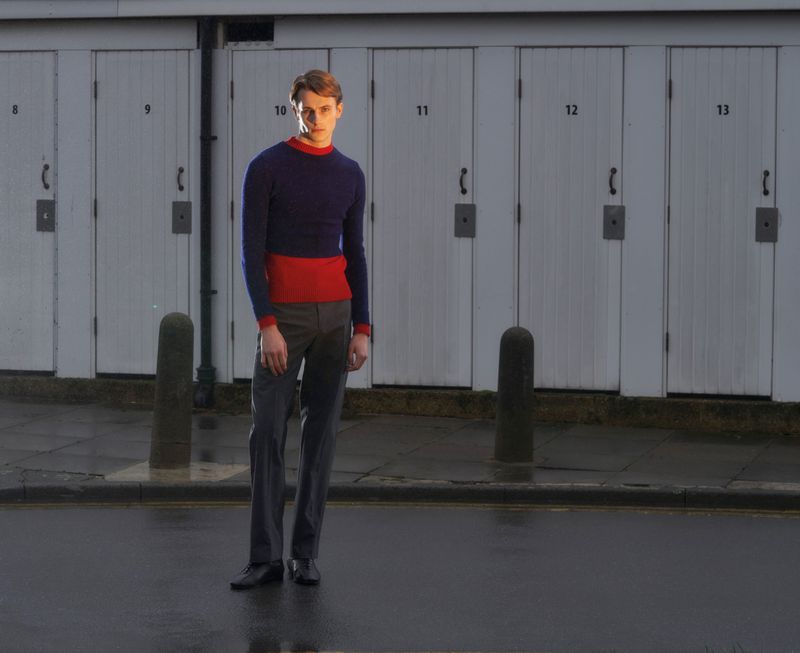 A young male model standing on an asphalt street in front of white paneled doors wearing a navy and red sweater with grey trousers.