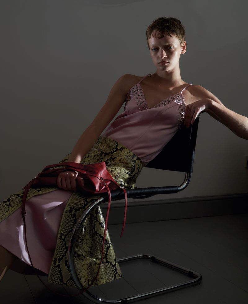 Model with short hair seated in a chair, wearing a pink top and snakeskin print skirt while holding a red handbag.
