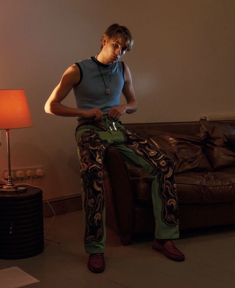Young male model seated on a leather sofa wearing a blue tank top and green patterned trousers in a dimly lit room.