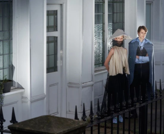Two models wearing avant-garde sculptural fashion standing in front of a white residential building exterior with a black metal fence.