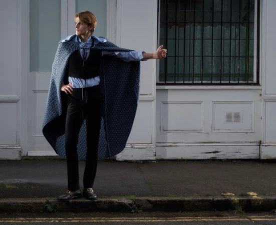 Model wearing a blue patterned cape over a black vest and trousers standing outside a white building