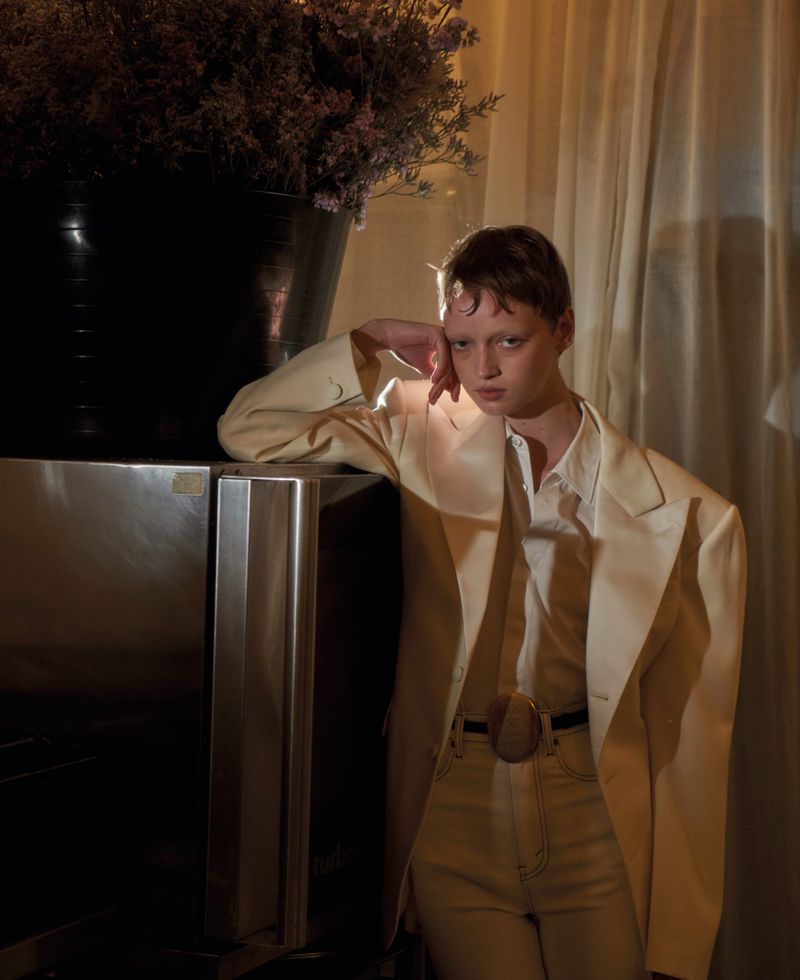 Model in cream blazer and white shirt leaning against a dark refrigerator with a large floral vase