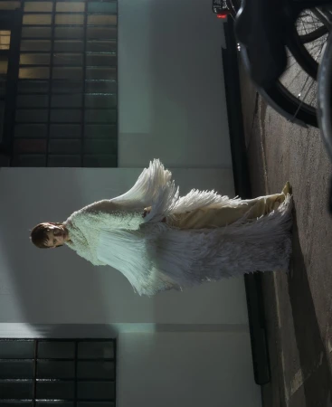 Model wearing a heavily fringed white textured garment standing outdoors against a building exterior wall.