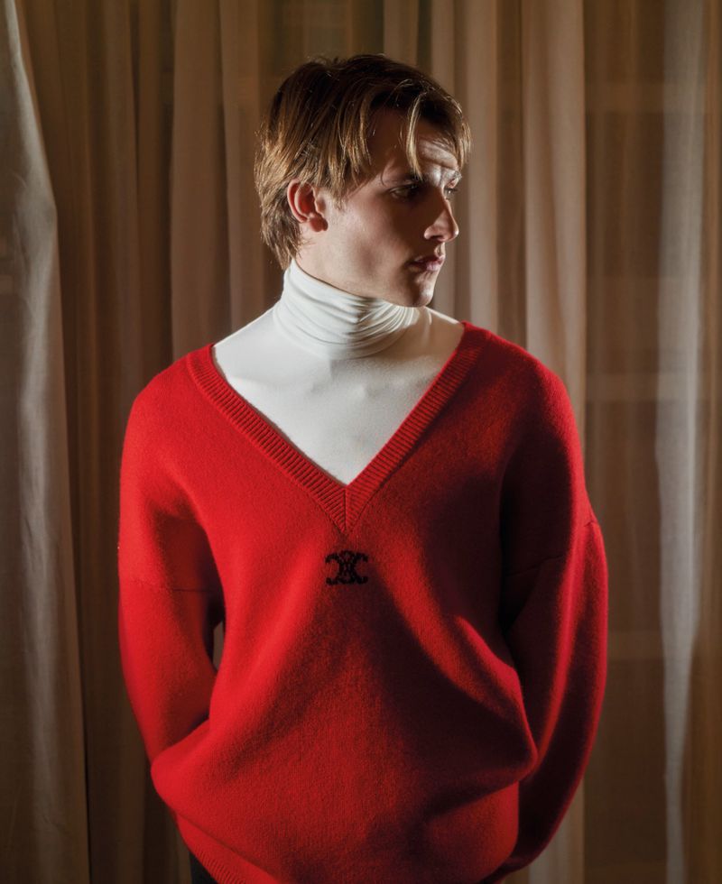 A young male model wearing a bright red V-neck sweater over a white turtleneck against draped fabric curtains.