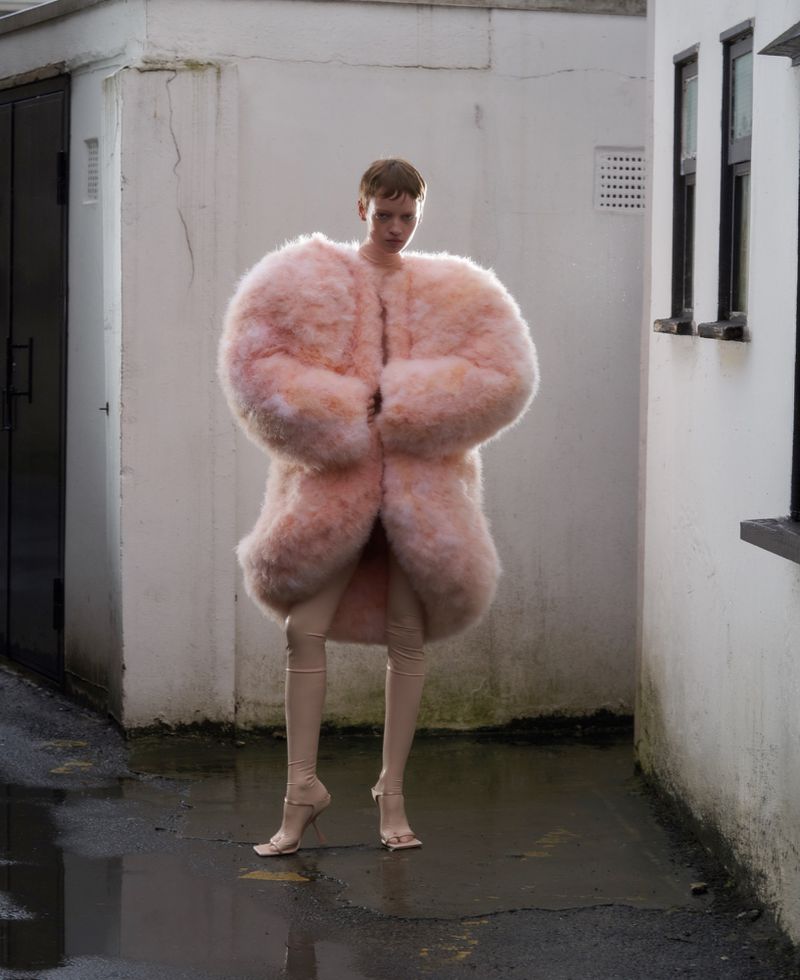 Model standing in an alleyway wearing a voluminous pink fuzzy coat and tall beige boots for ODDA Magazine