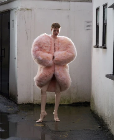 Model standing in an alleyway wearing a voluminous pink fuzzy coat and tall beige boots for ODDA Magazine
