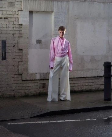 Model in a pink blouse and white wide-leg trousers standing on a city street beside a brick building exterior.