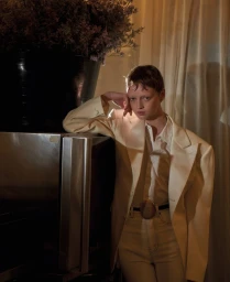 Model in cream blazer and white shirt leaning against a dark refrigerator with a large floral vase