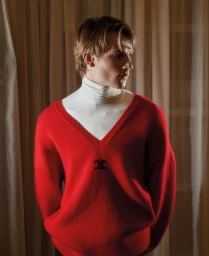 A young male model wearing a bright red V-neck sweater over a white turtleneck against draped fabric curtains.