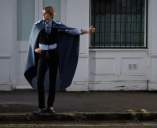 Model wearing a blue patterned cape over a black vest and trousers standing outside a white building